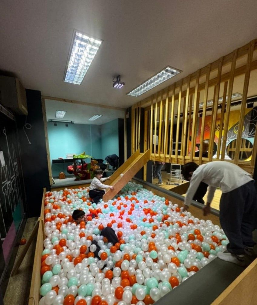 Niños jugando en una piscina de pelotas con pelotas blancas, naranjas y verde azulado. Una se desliza por una pequeña rampa.