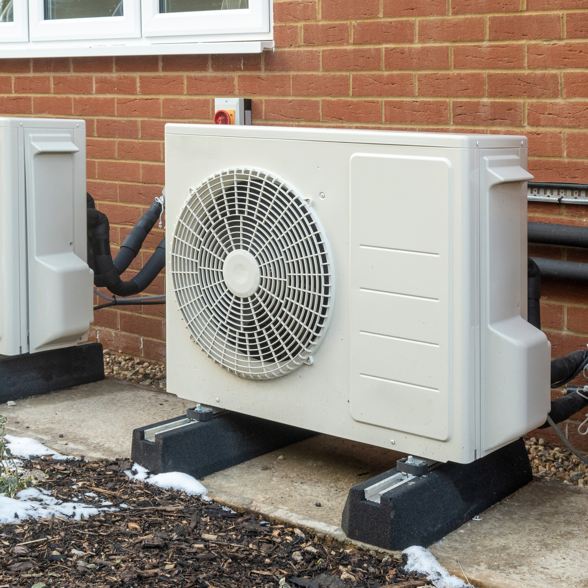 Outdoor air conditioning unit beside a brick house, mounted on black supports with ductwork attached