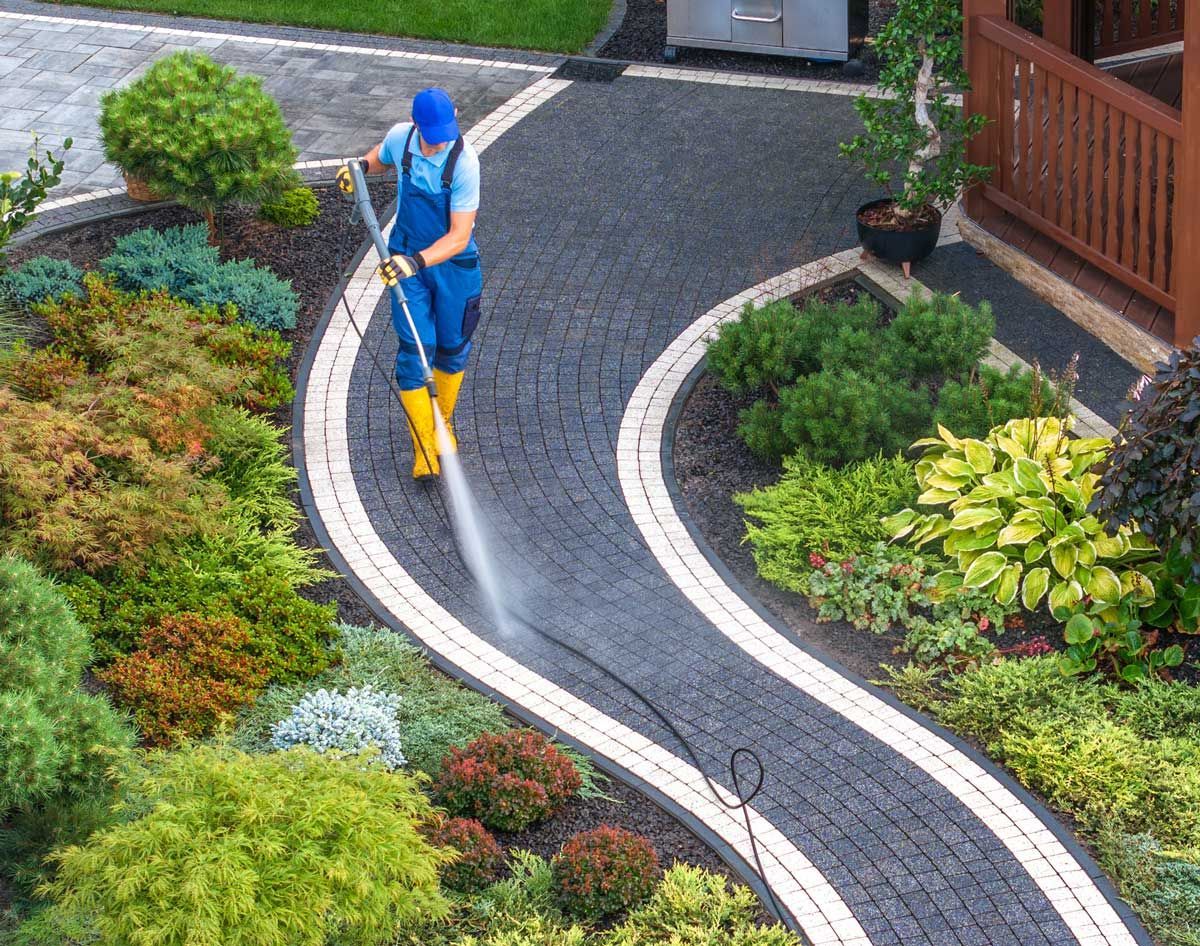 A man is using a high pressure washer to clean a brick walkway in a garden.