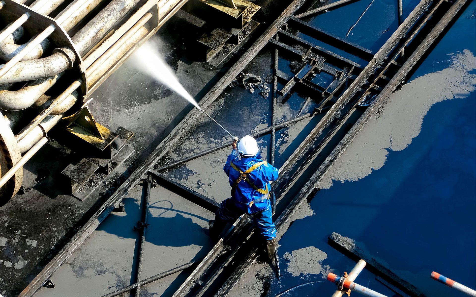 Worker in blue suit spraying water on a dark, oil-covered structure, likely a ship deck.