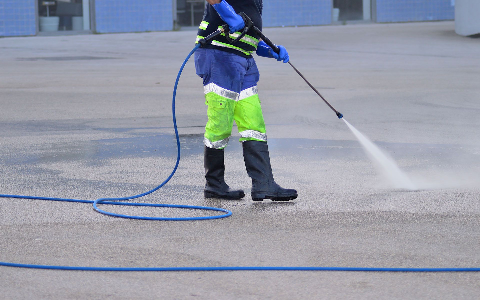 Person in protective gear pressure washes a concrete surface with a water hose.