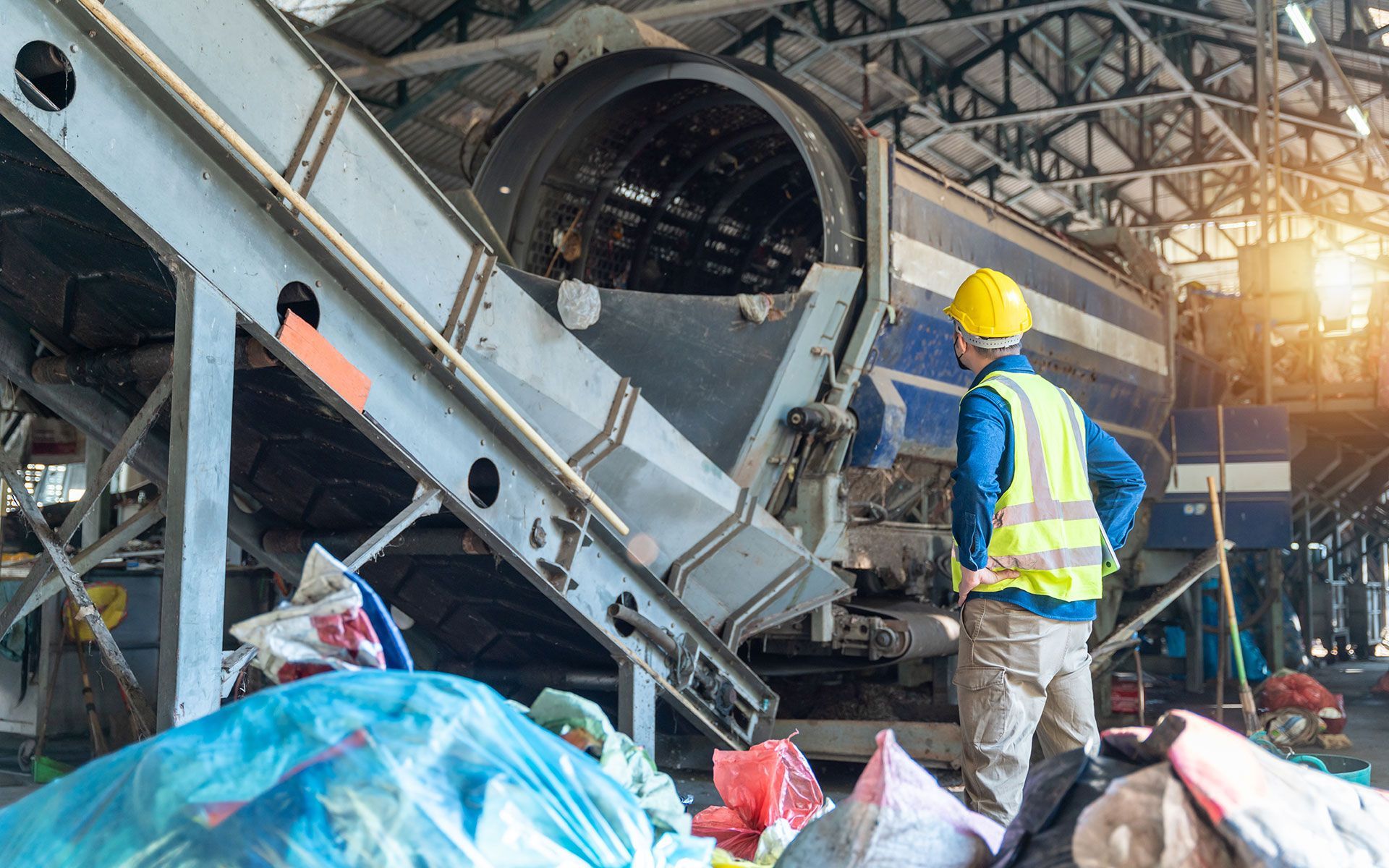Worker in a yellow hard hat and vest at a recycling facility, observing machinery and piles of sorted materials.