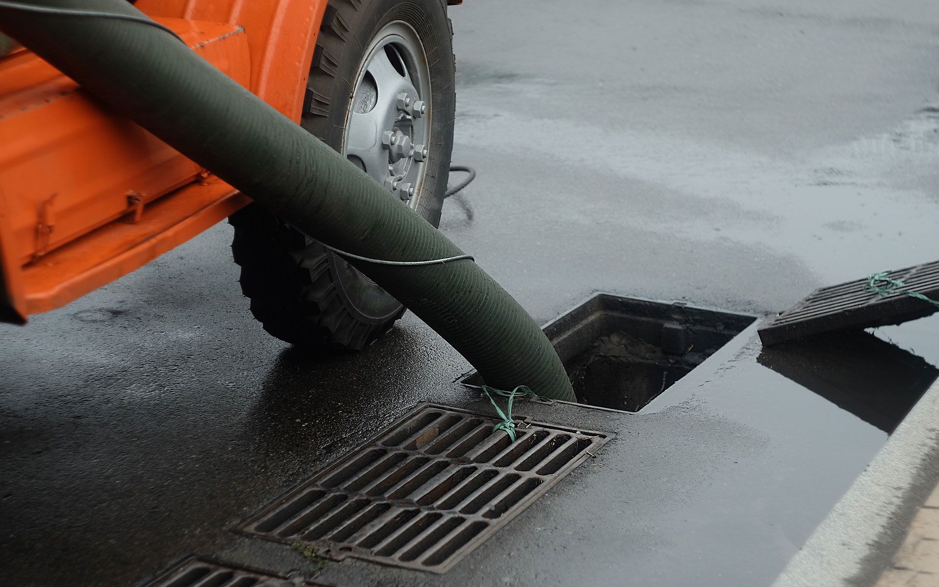 Orange truck hose draining water into a street drain on a wet asphalt surface.