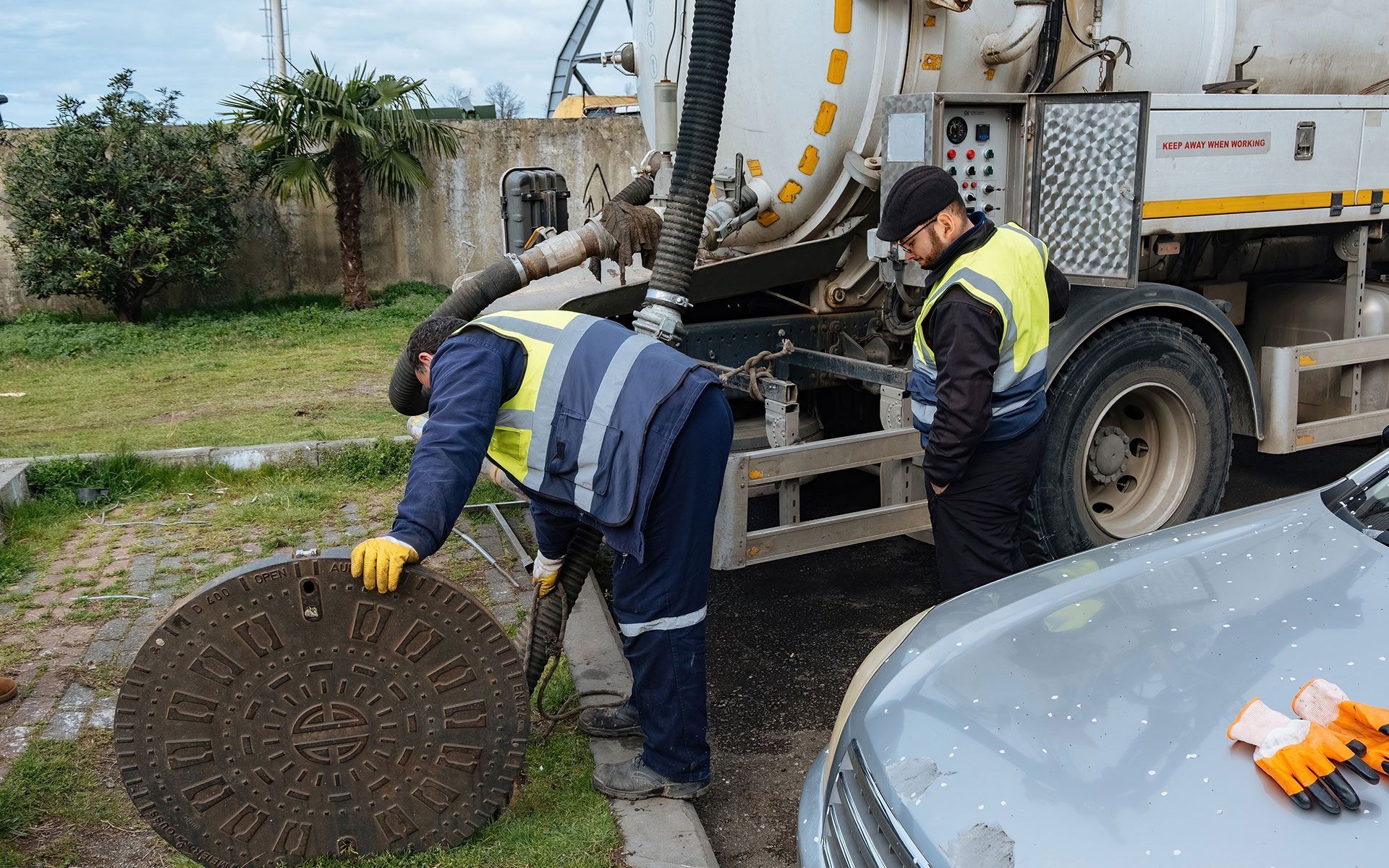 Two workers in vests tending to a manhole with a vacuum truck nearby.