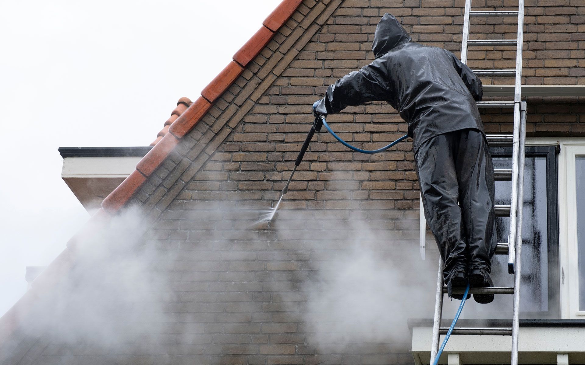 Person on ladder pressure washing brick house exterior, creating steam.