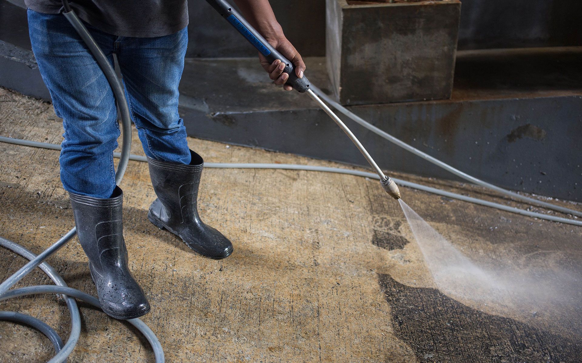 Person in jeans and boots power washing a concrete surface with a spray of water.
