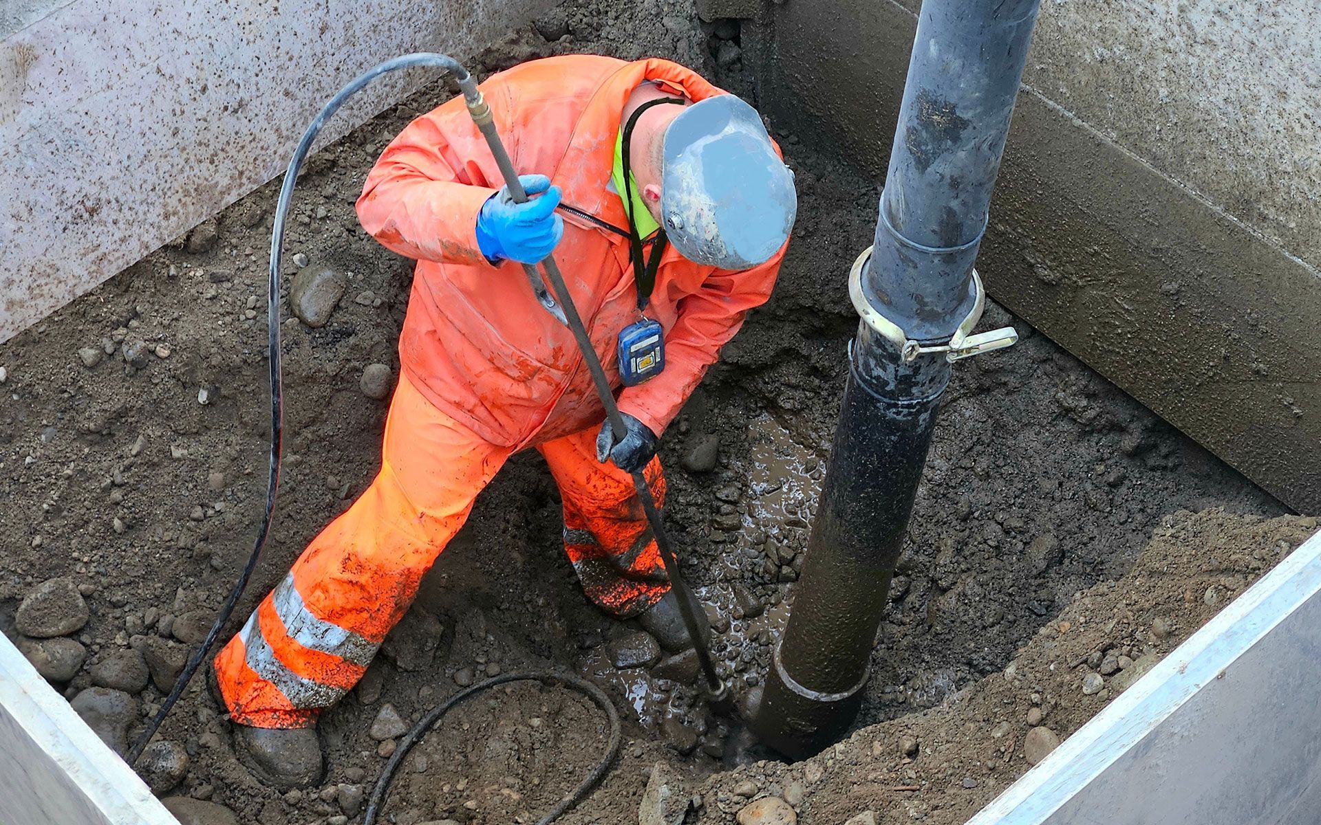 Worker in orange coveralls excavating around a pipe in a concrete pit.