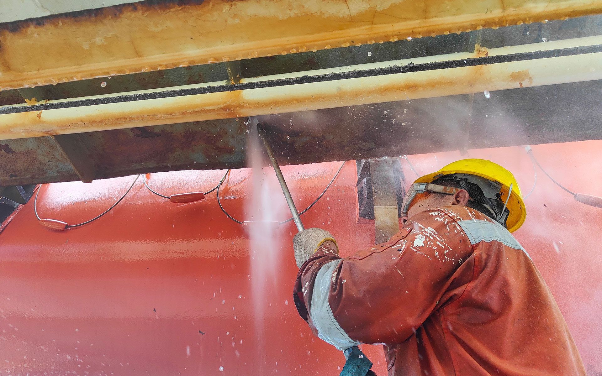 Man in protective gear using high-pressure water to clean a rusty metal beam on a red surface.