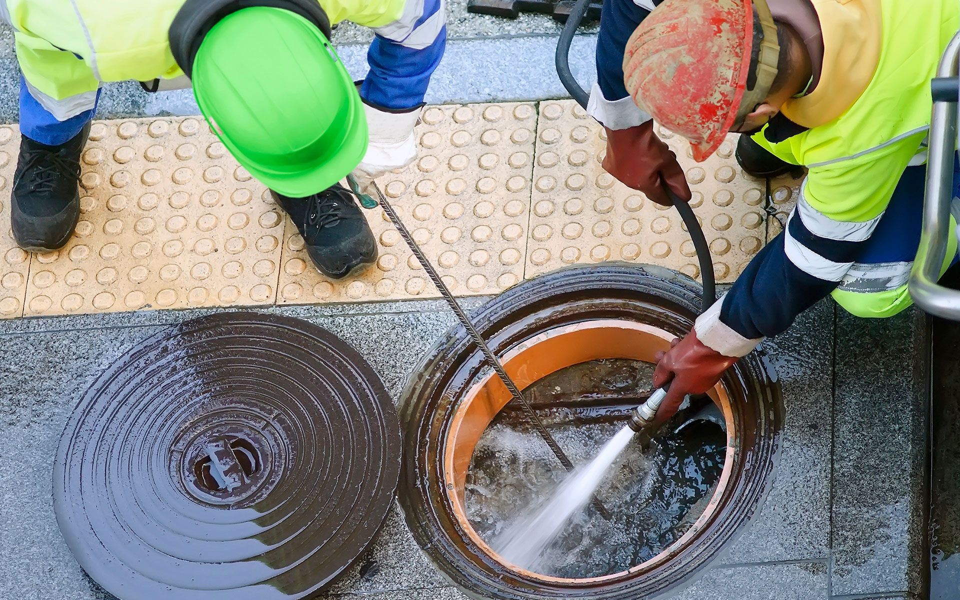 Workers cleaning a sewer drain with high-pressure water. One wears a green hard hat, the other a red one.