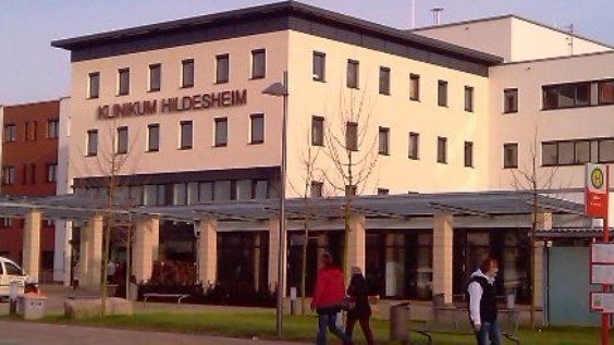 Außenansicht des Klinikums Hildesheim mit Passanten und einer Bushaltestelle. Beiges Gebäude, braune Markise, bewölkter Himmel.
