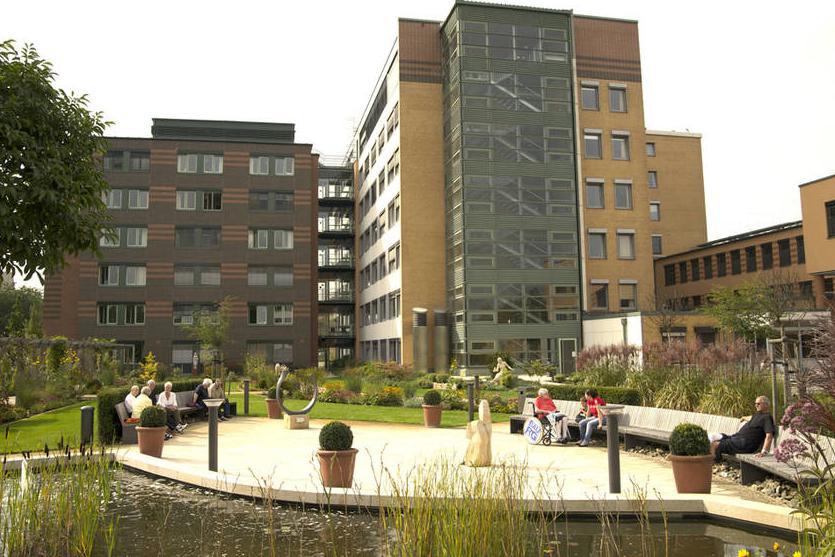 Außenansicht eines mehrstöckigen Gebäudes mit Garten, Wasserspiel und auf Bänken sitzenden Personen.