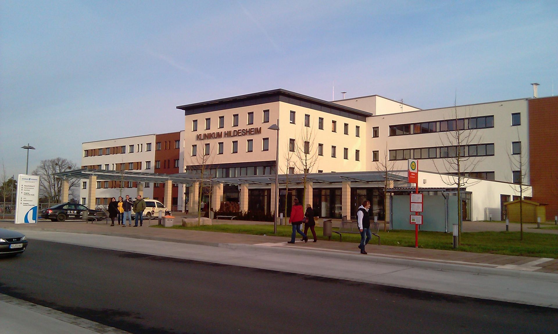 Außenansicht des Krankenhauses mit Fußgängern, Autos und einer Bushaltestelle.