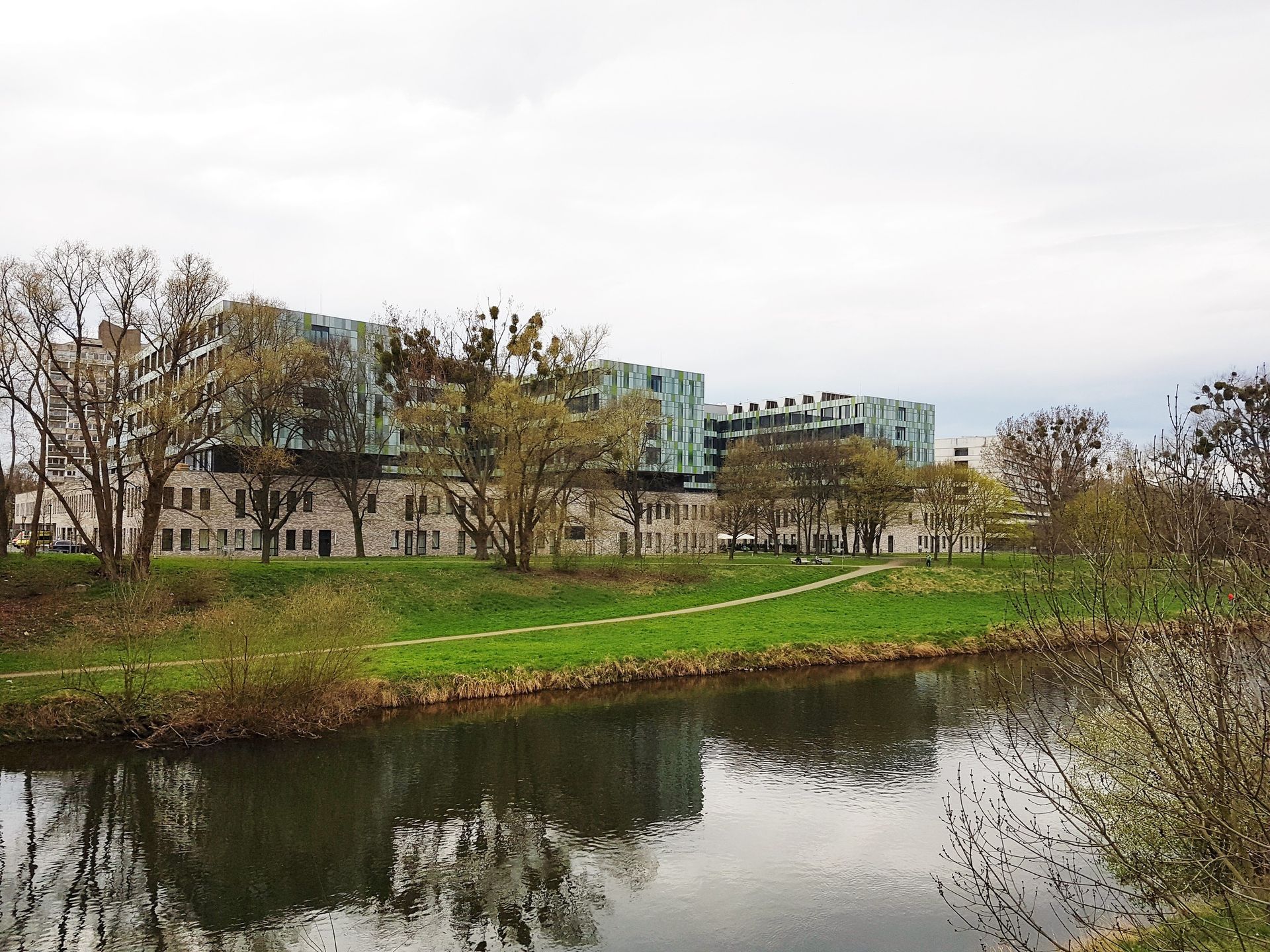 Ein Fluss mit grünen Ufern, Bäumen und modernen Gebäuden unter bedecktem Himmel.