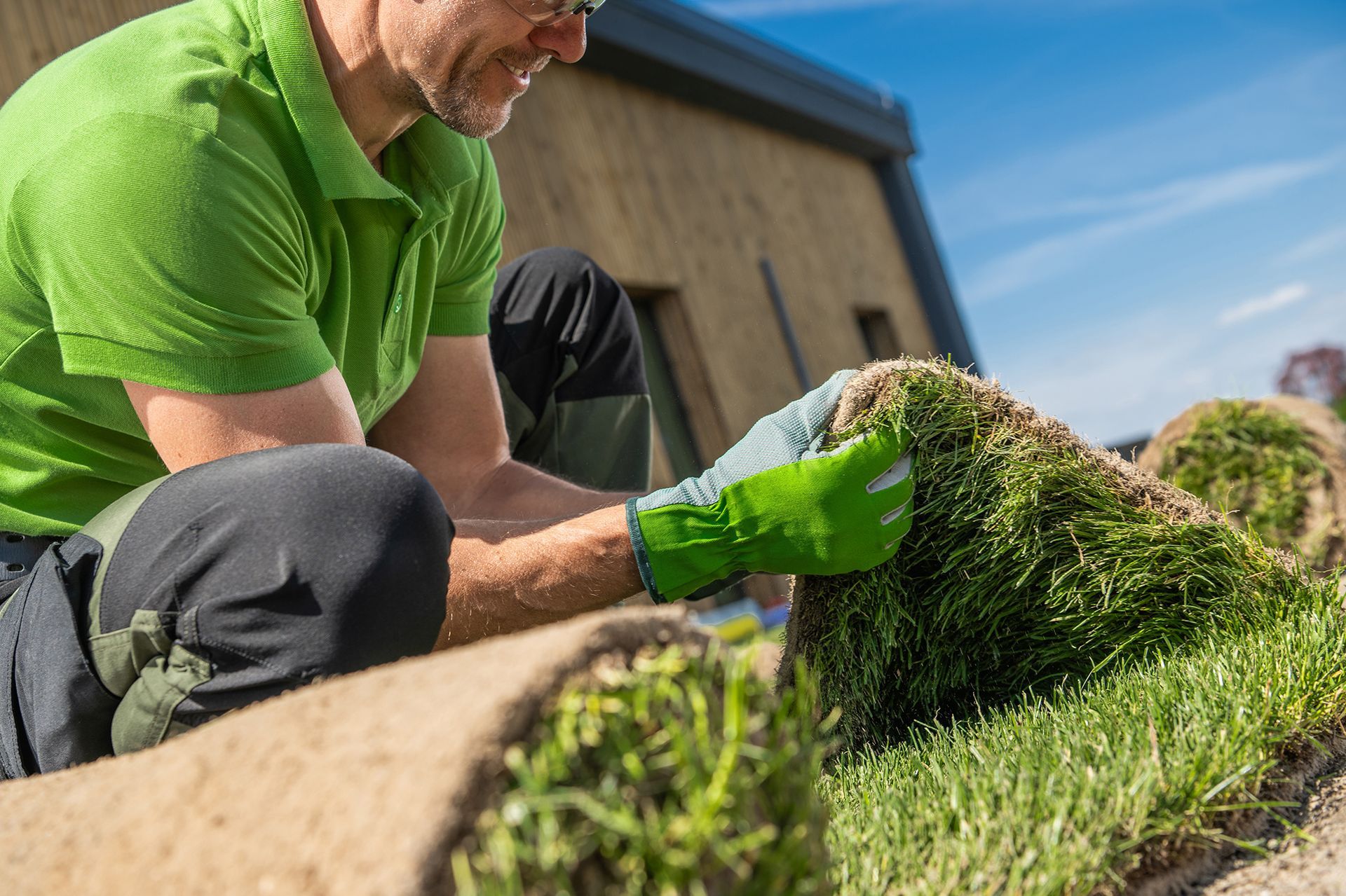 A man in a green shirt and green gloves is rolling a roll of grass.