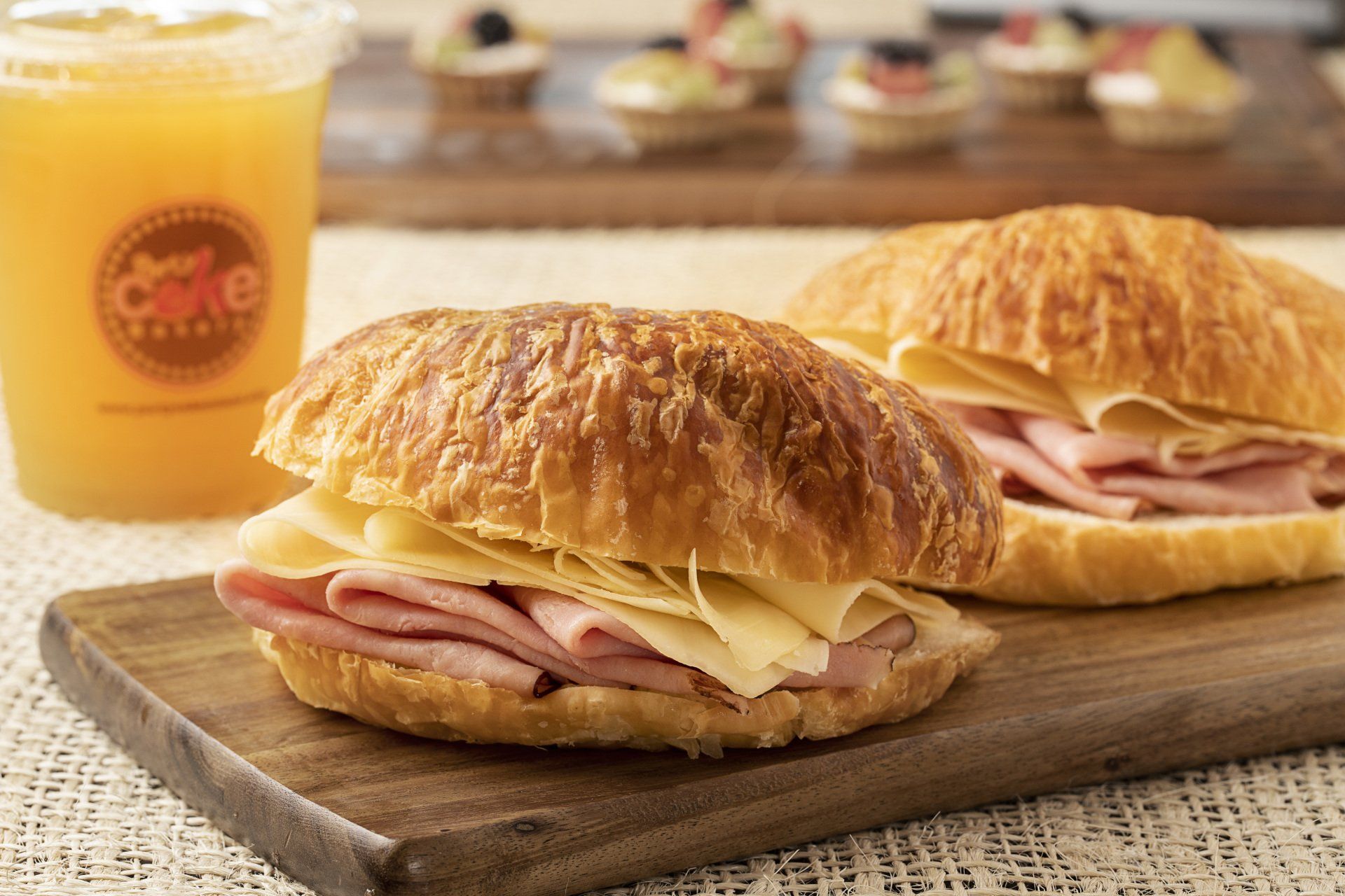 Ham and cheese croissant sandwiches on a wooden board, with orange juice and pastries in the background.