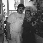 Man in white t-shirt with apron and woman arm-in-arm, standing near a window with sunflowers.