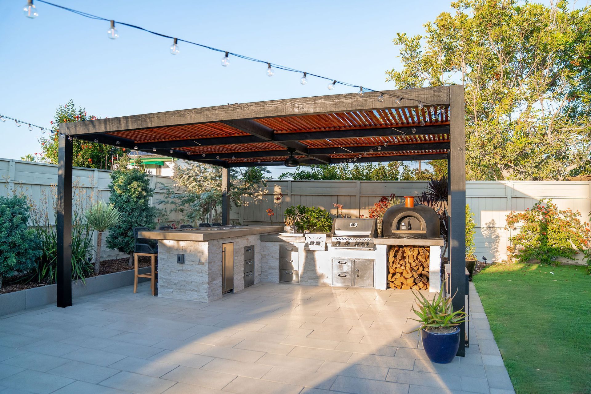 Outdoor kitchen with a pizza oven, grill, and stone countertop under a wood-slatted pergola on a paved patio.