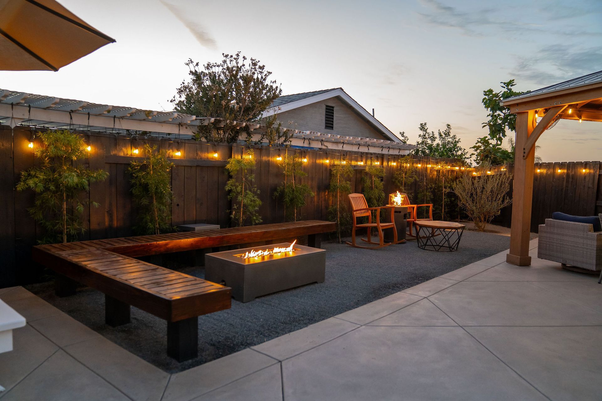 Backyard patio with fire pit, wooden benches, and string lights, at dusk.