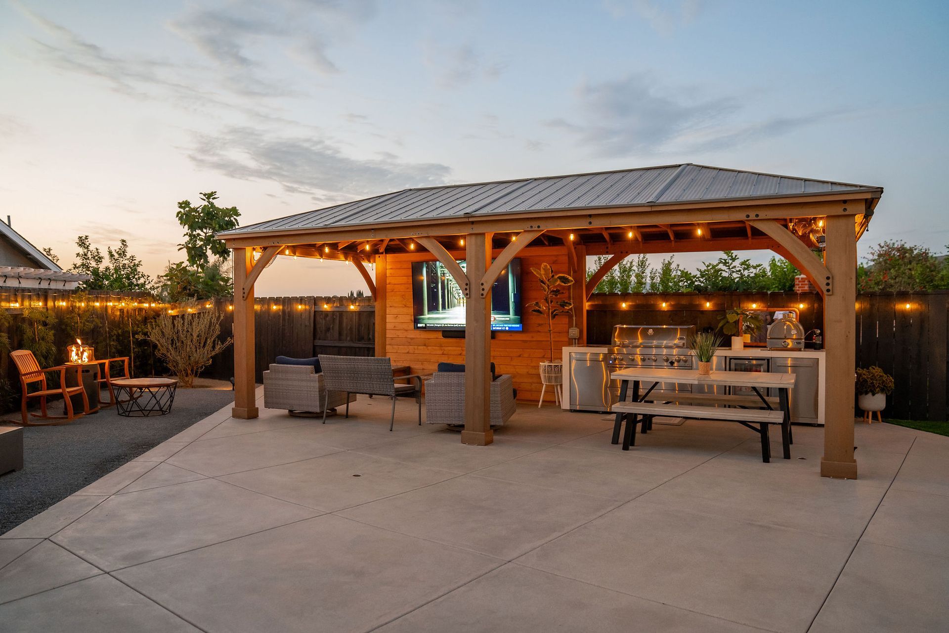 A backyard patio with a wooden gazebo, an outdoor TV, dining area, grill, and string lights at sunset.