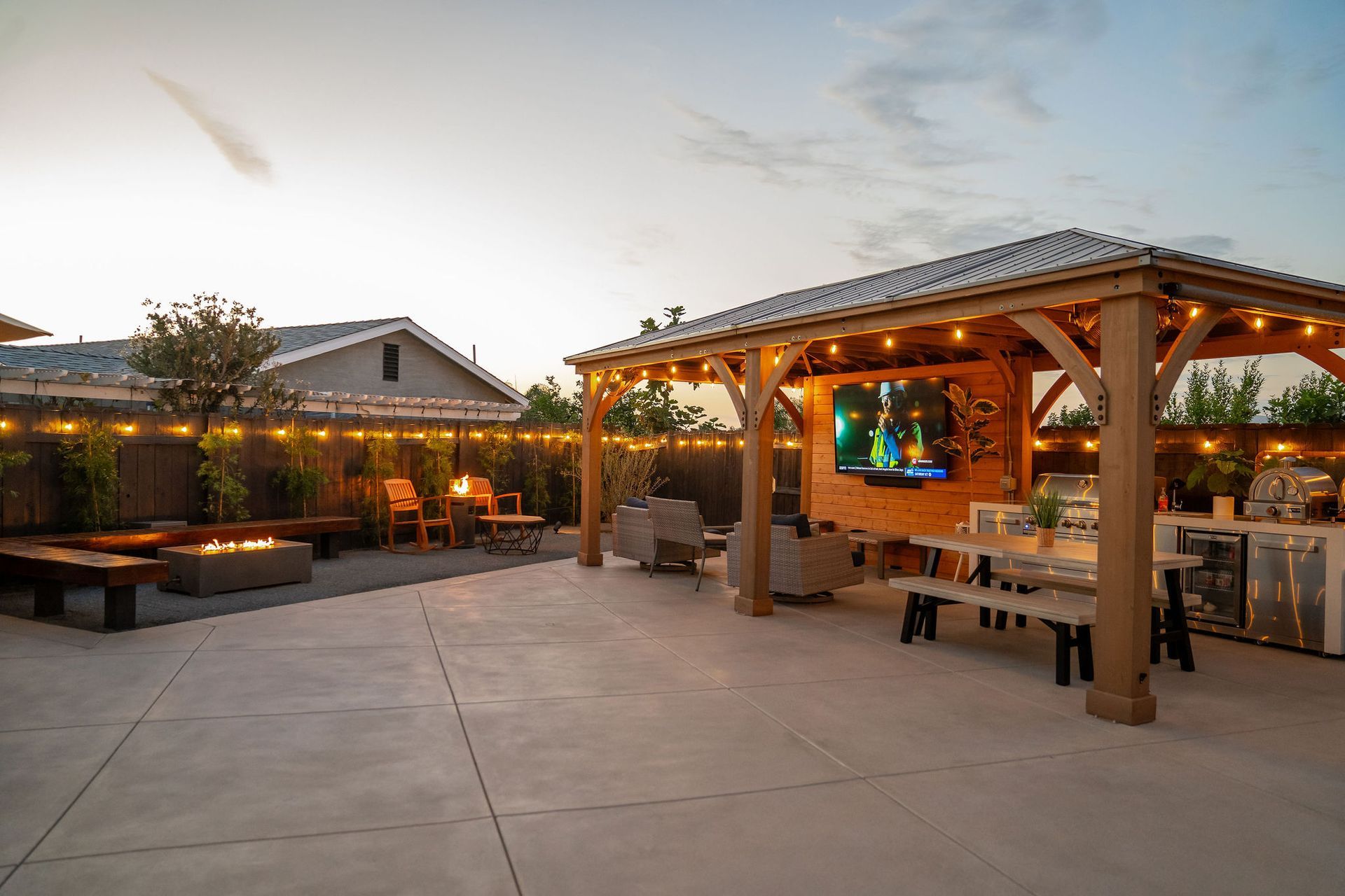 A backyard patio features a wooden pavilion with a wall-mounted TV, dining area, and a separate fire pit seating lounge.