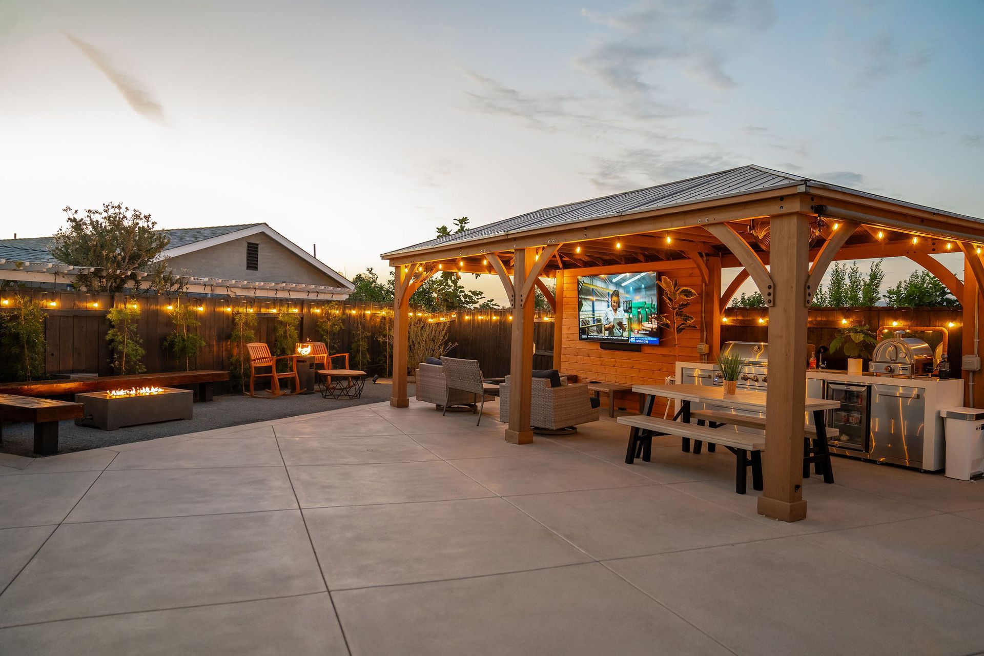 Outdoor patio with a wooden gazebo, an entertainment area featuring a TV, a dining set, and a fire pit at dusk.