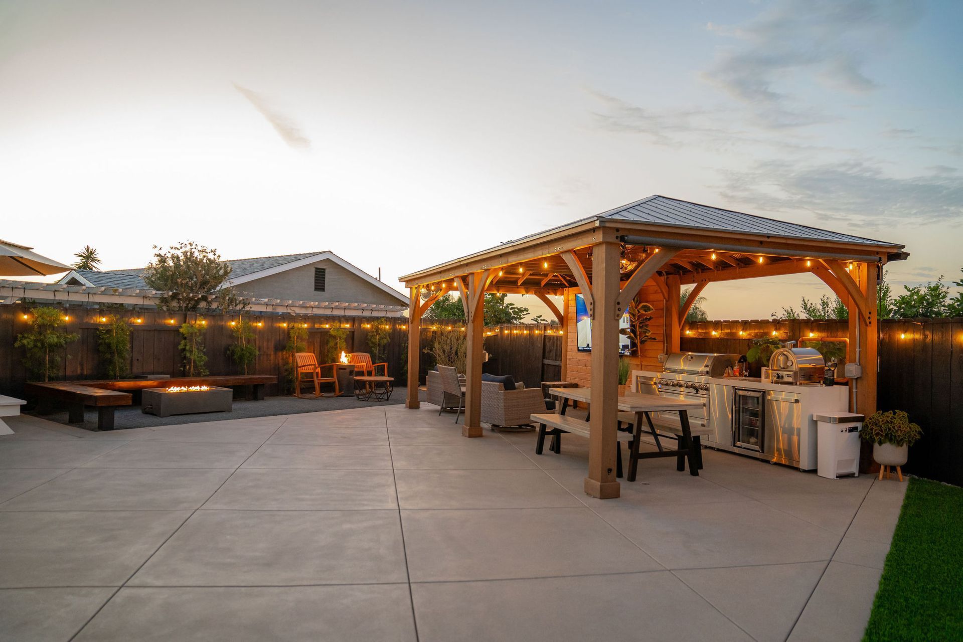 A sunset view of a backyard patio with a wooden gazebo, outdoor kitchen, picnic table, and a fire pit area.