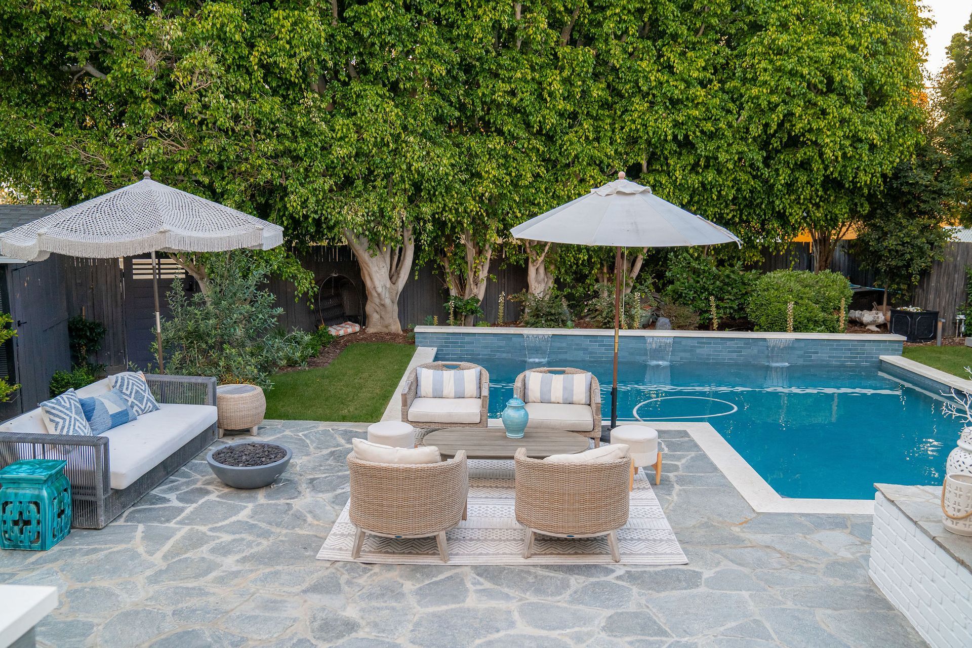 Patio with seating around a pool; gray stone, white umbrellas, lush green trees.