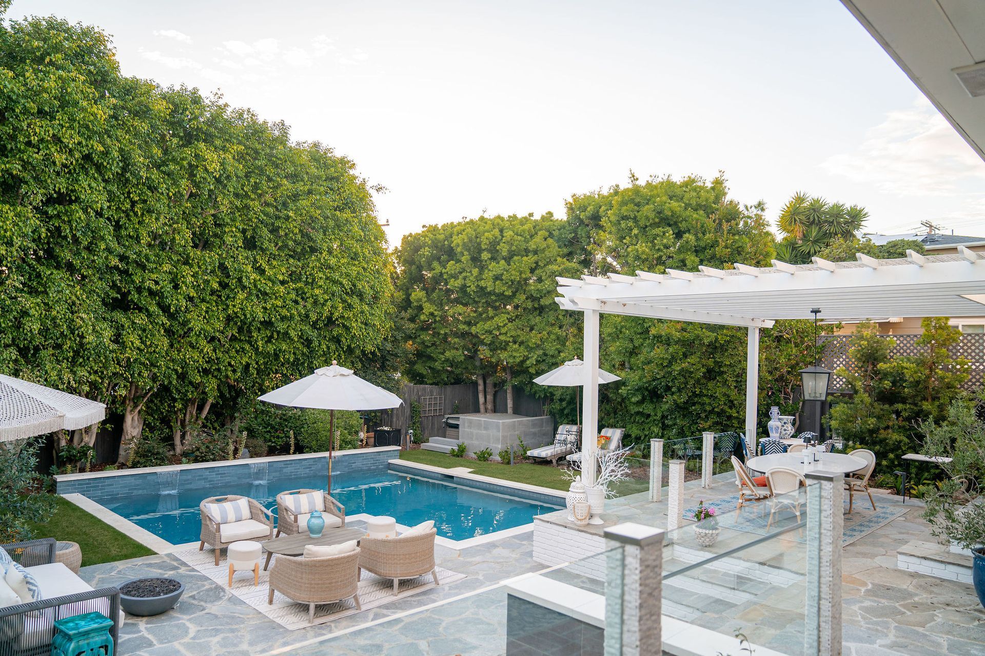 A sunny backyard with a rectangular pool, light-colored patio furniture, a white pergola, and lush green trees.
