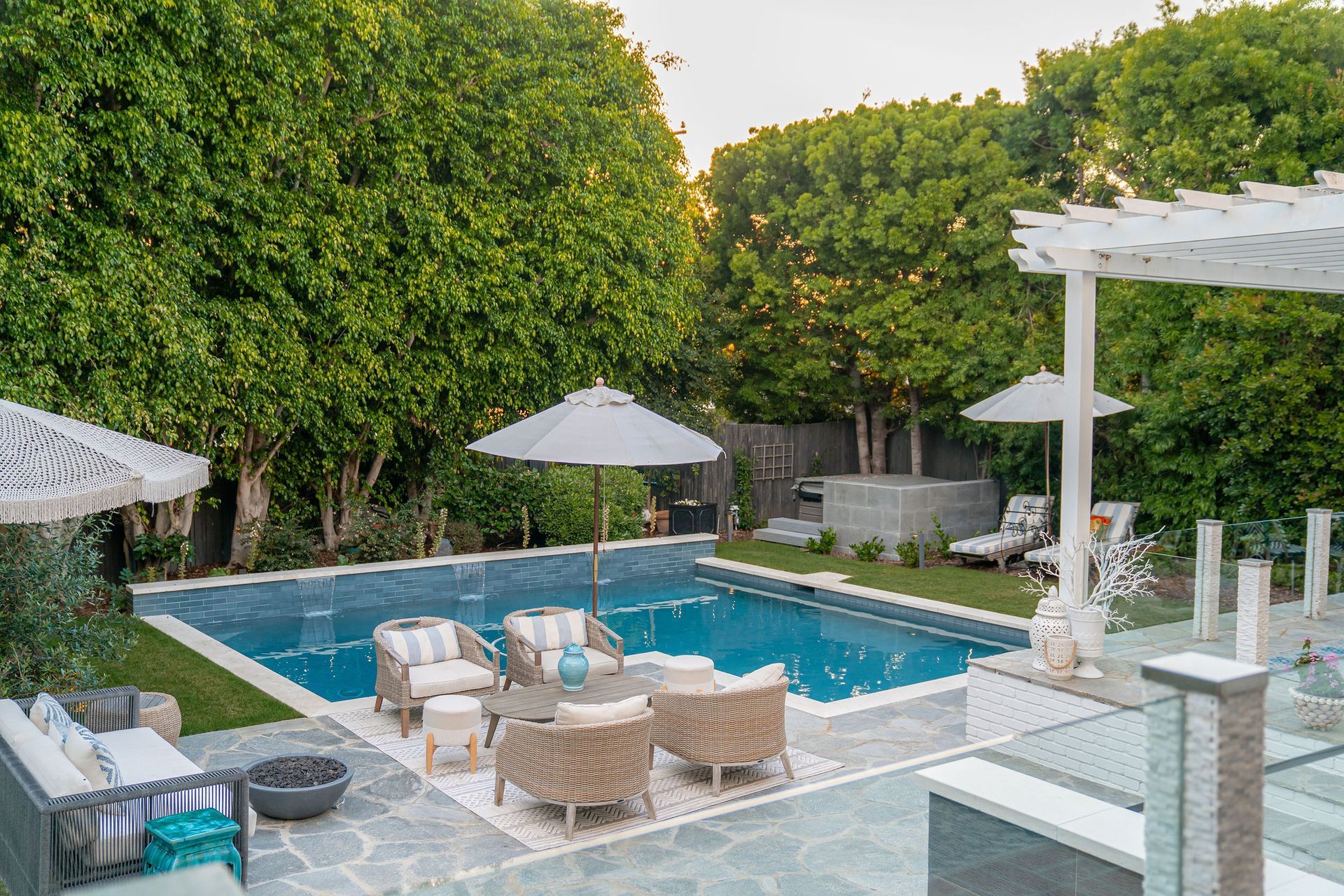 A sunny patio with a swimming pool, lounge chairs, umbrellas, and lush greenery in a backyard setting.