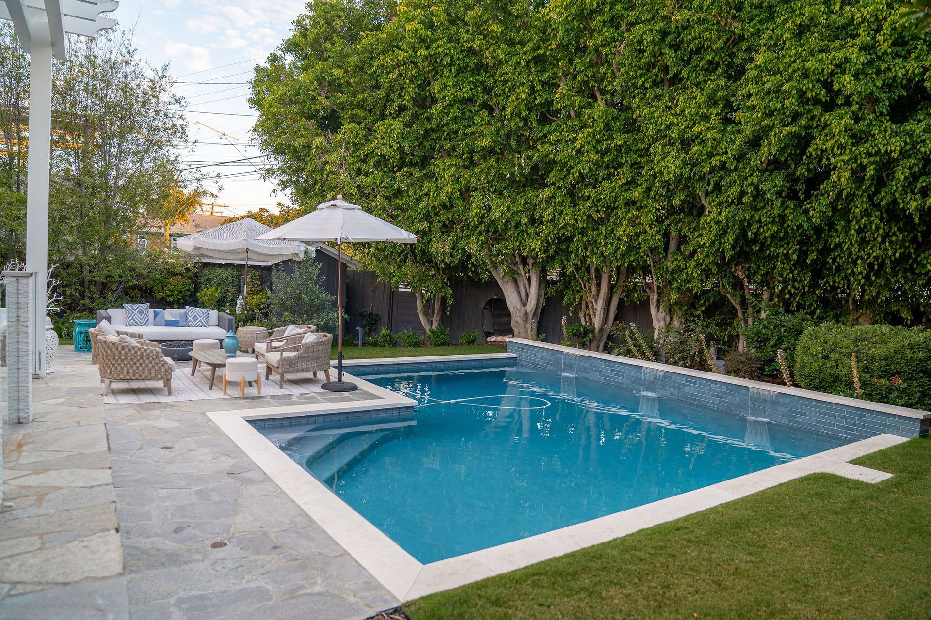 A sunlit backyard with a swimming pool, lounge furniture under white umbrellas, and large trees in the background.