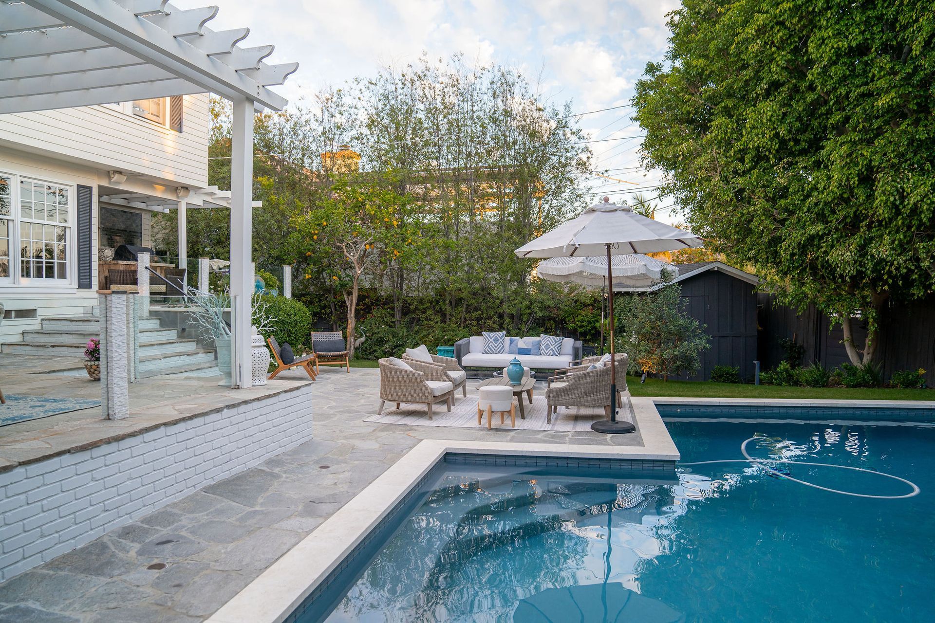 A blue swimming pool sits beside a stone patio with outdoor lounge furniture under an umbrella, near a house with a pergola.