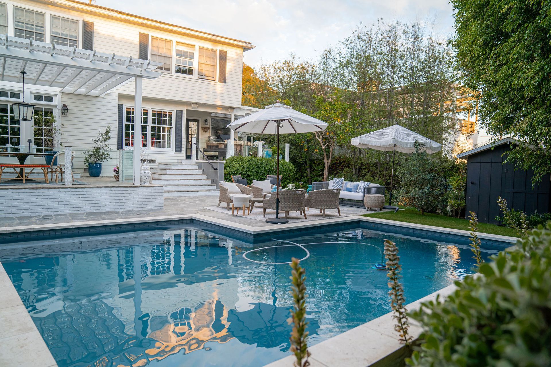 A white multi-story house with a backyard pool, patio furniture, and white umbrellas under a clear, sunny sky.