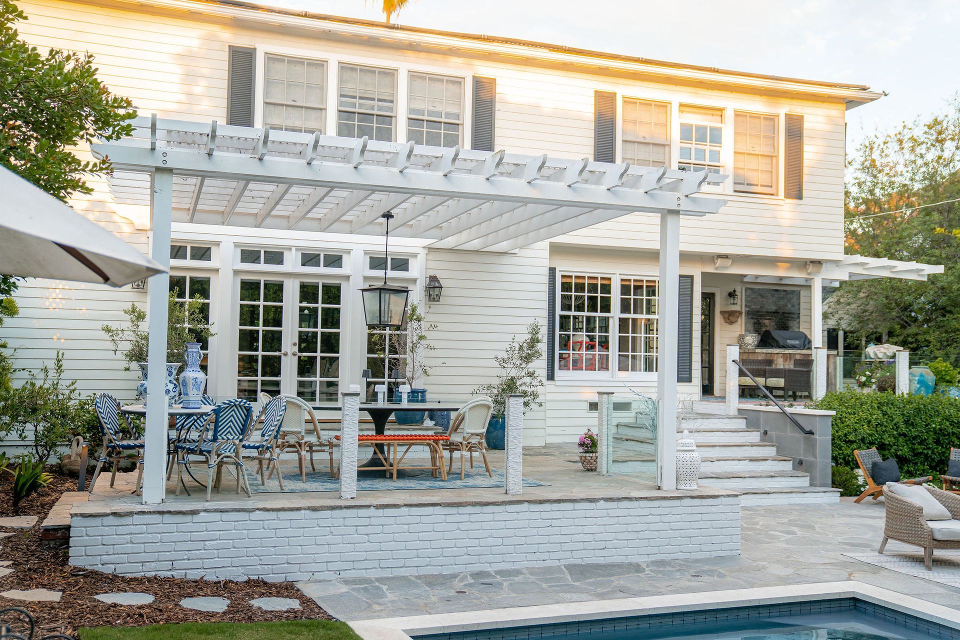 A white two-story house with a pergola-covered stone patio, outdoor dining area, and a swimming pool in the backyard.