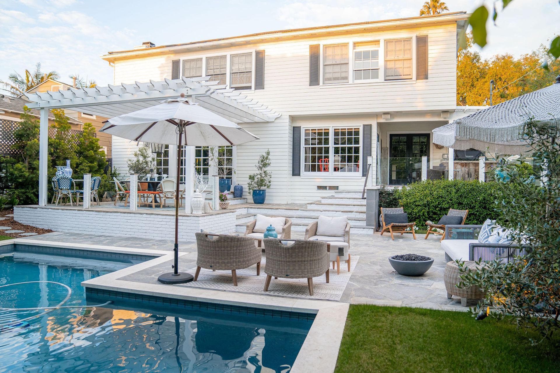 Backyard with pool, patio, and white two-story house. Patio furniture includes chairs, table, and umbrella. Lush green grass.