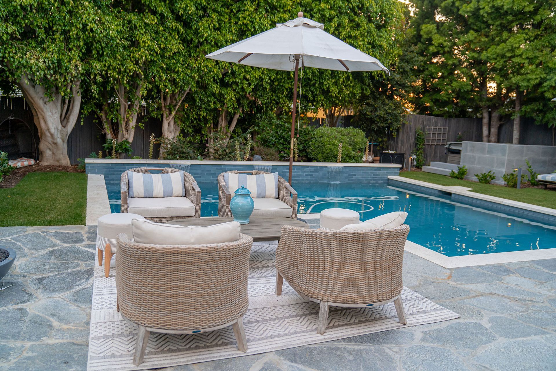 Four woven chairs surround a small table on a patio by a pool, under a white umbrella, with trees in the background.