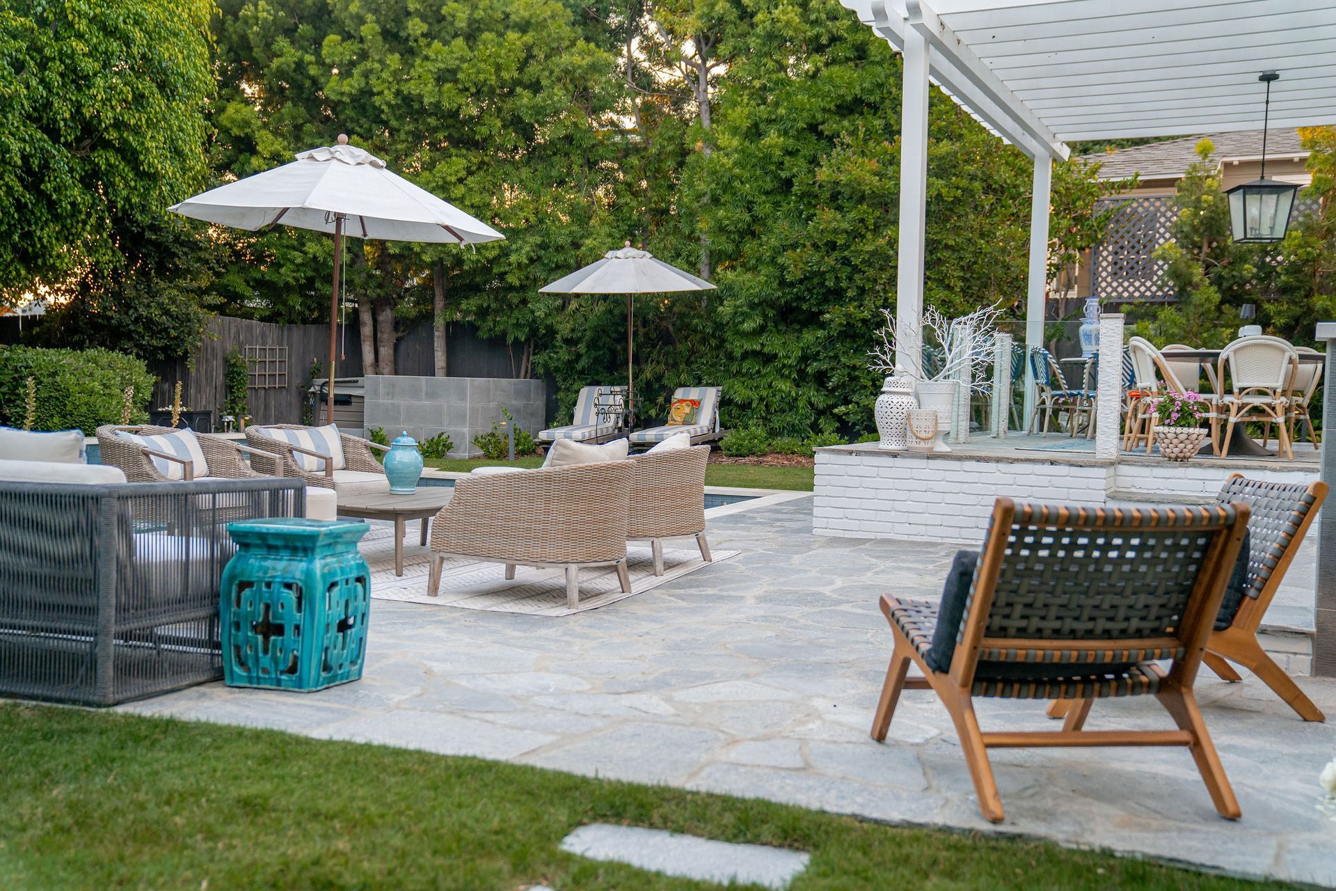 A patio with a pergola, stone flooring, multiple chairs, umbrellas, and a bright teal garden stool surrounded by trees.