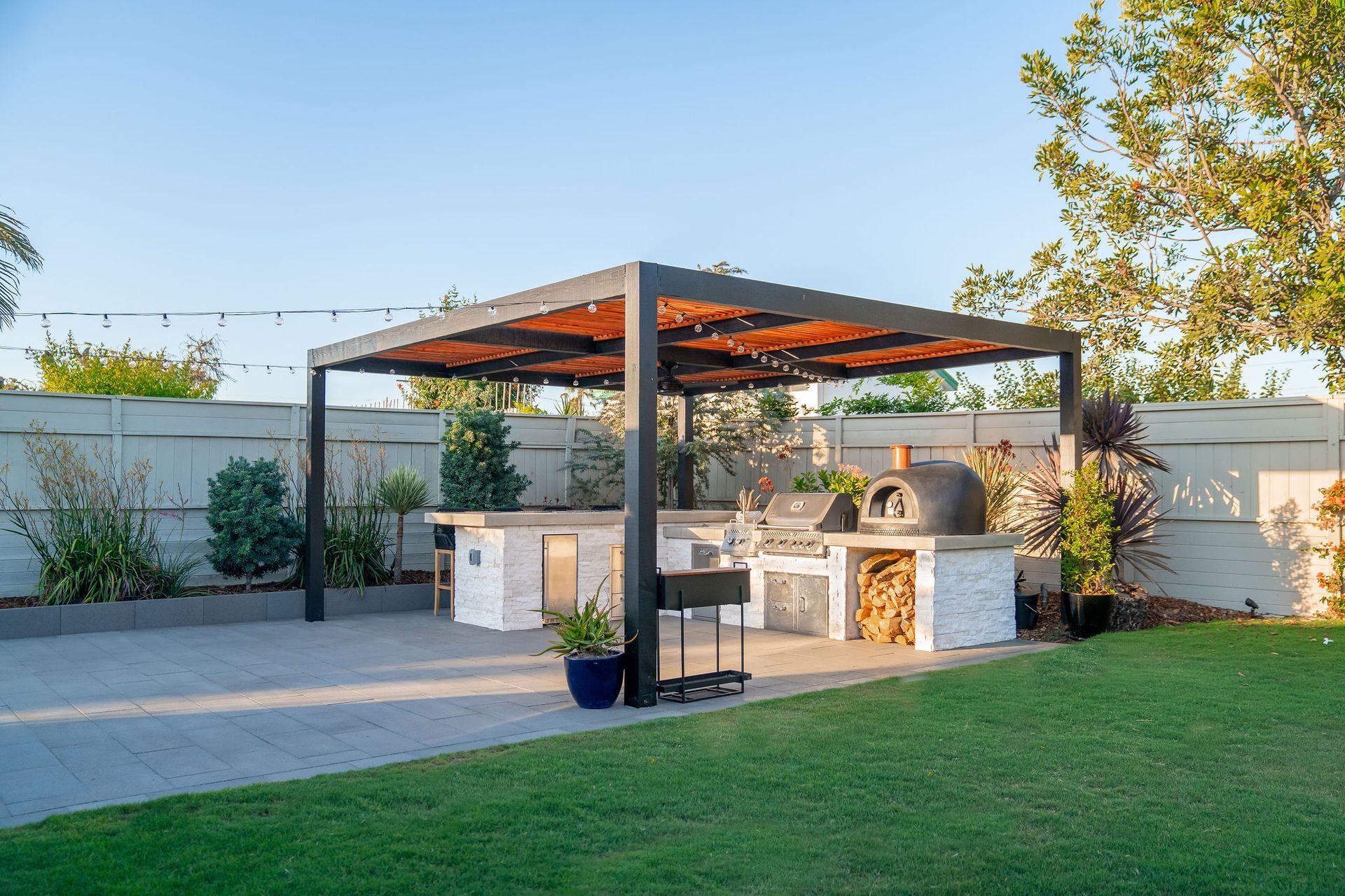 Outdoor kitchen with pergola, stone counters, grill, pizza oven, and green lawn.