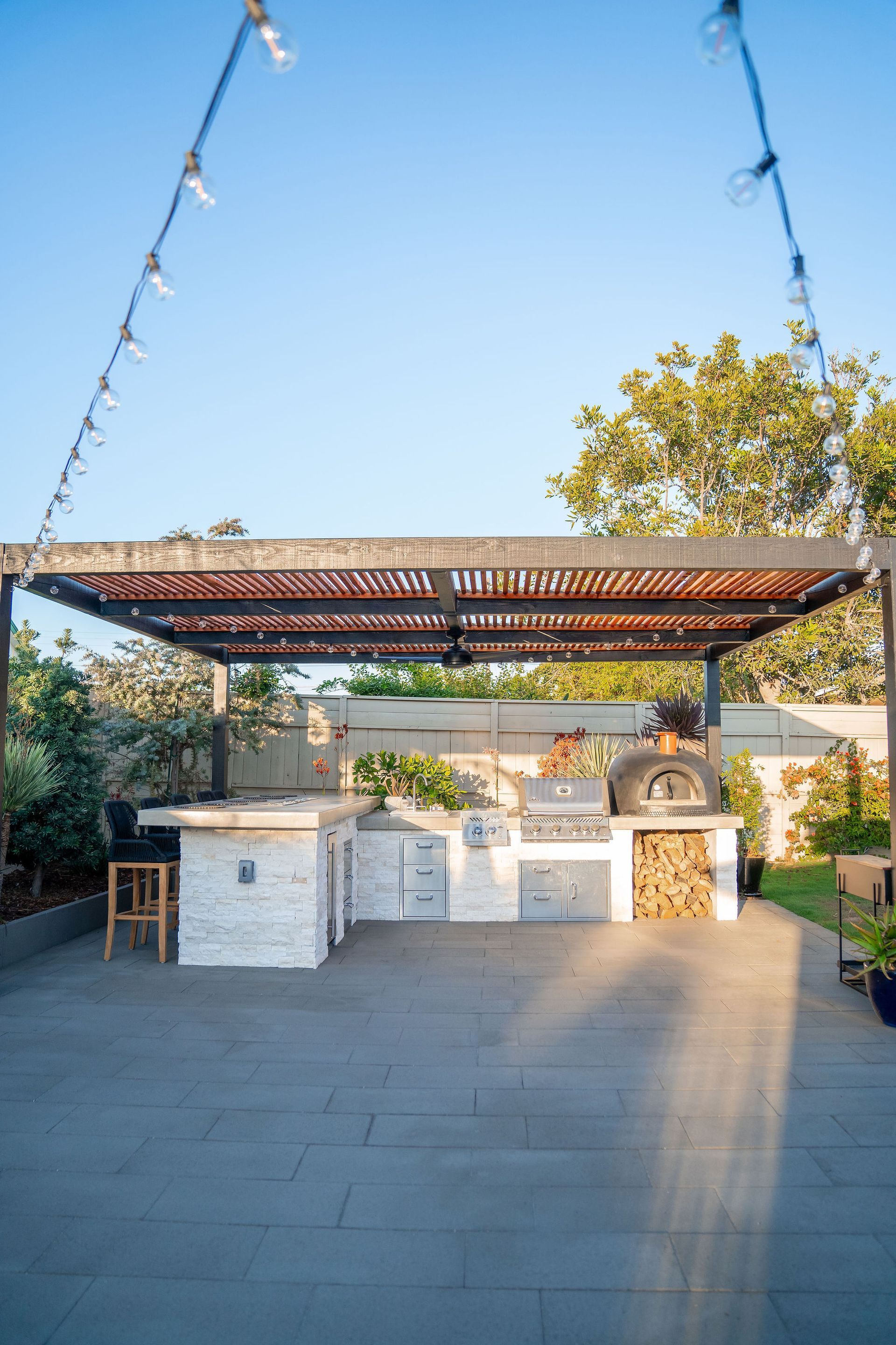 A modern outdoor kitchen with a stone counter, grill, and pizza oven under a wooden pergola, illuminated by string lights.