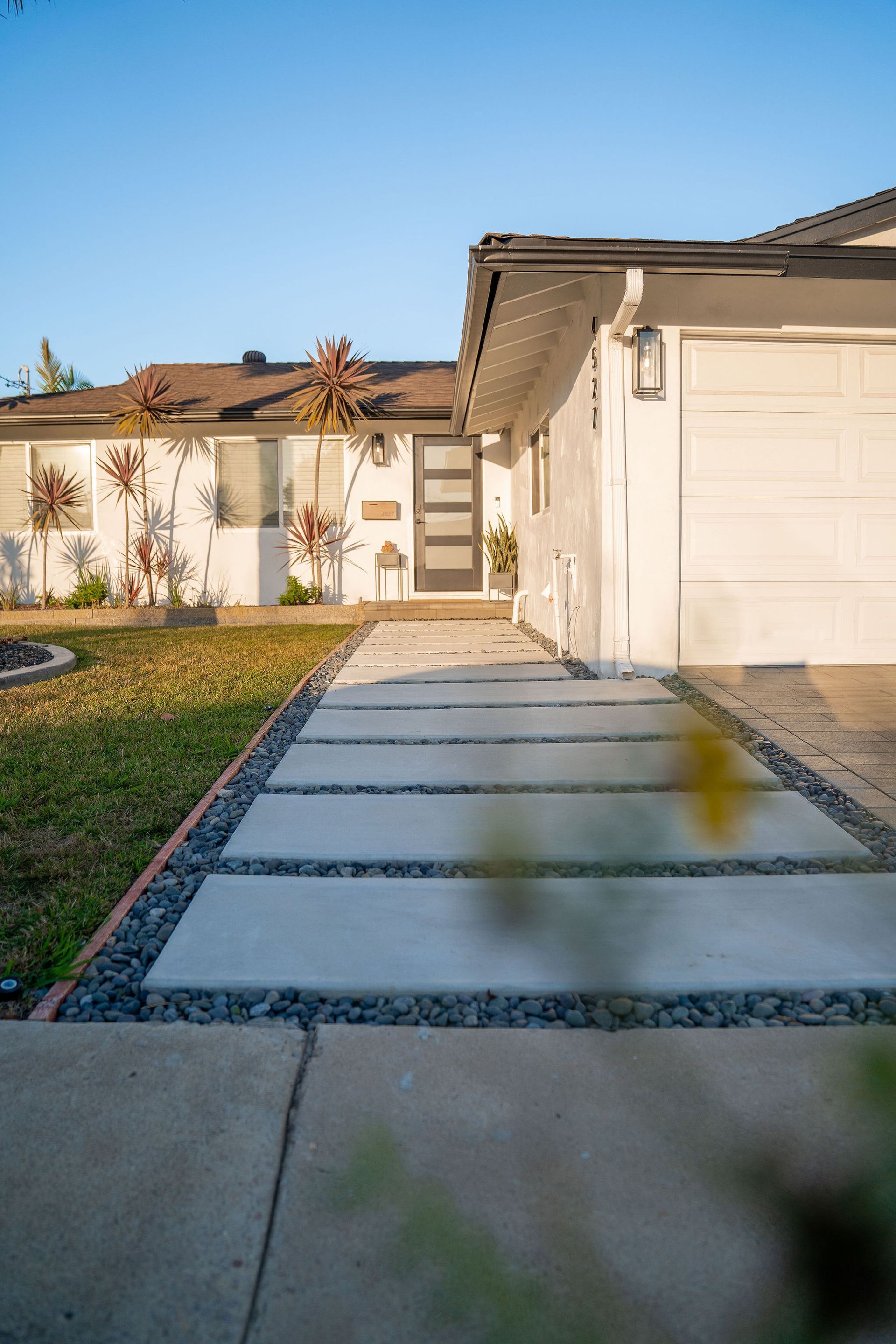 A modern concrete paver walkway leads to a white house under a clear blue sky, framed by dark gravel and a grassy lawn.