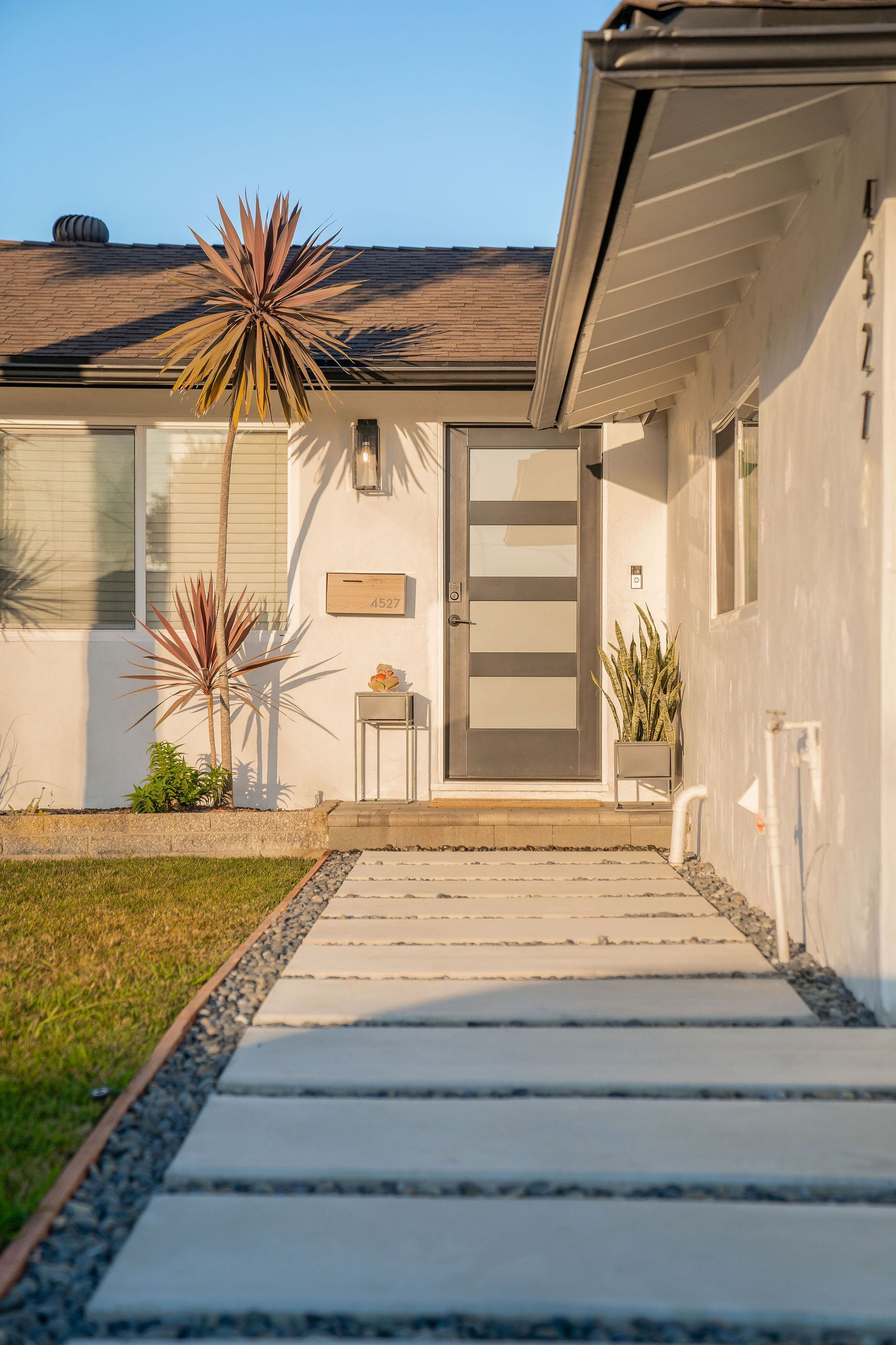 Modern house entrance with a gray front door, light stucco walls, and a concrete paver walkway leading to the porch.