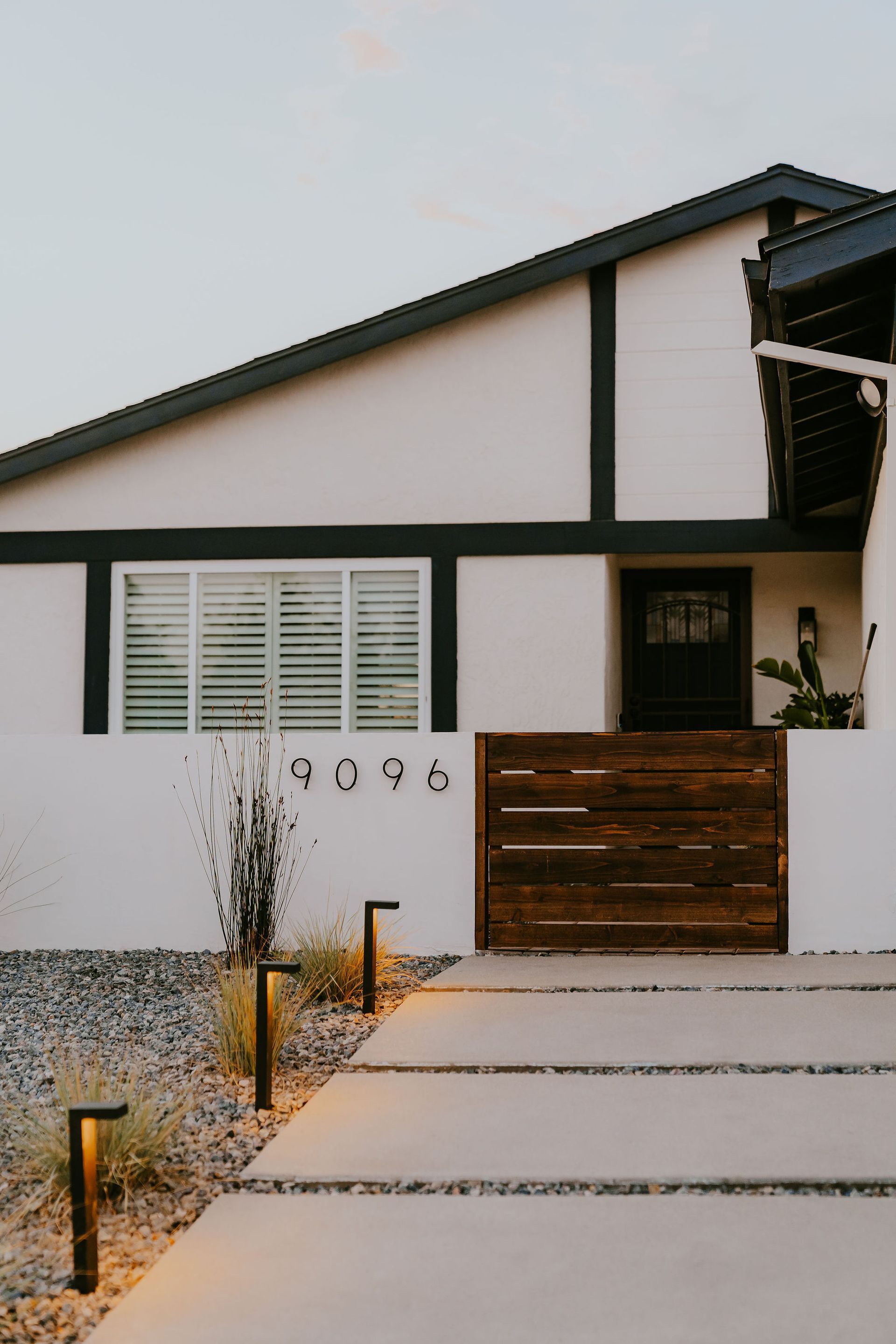 A modern house with white stucco walls, dark wood trim, a wooden privacy gate, and a stone walkway with landscape lighting.