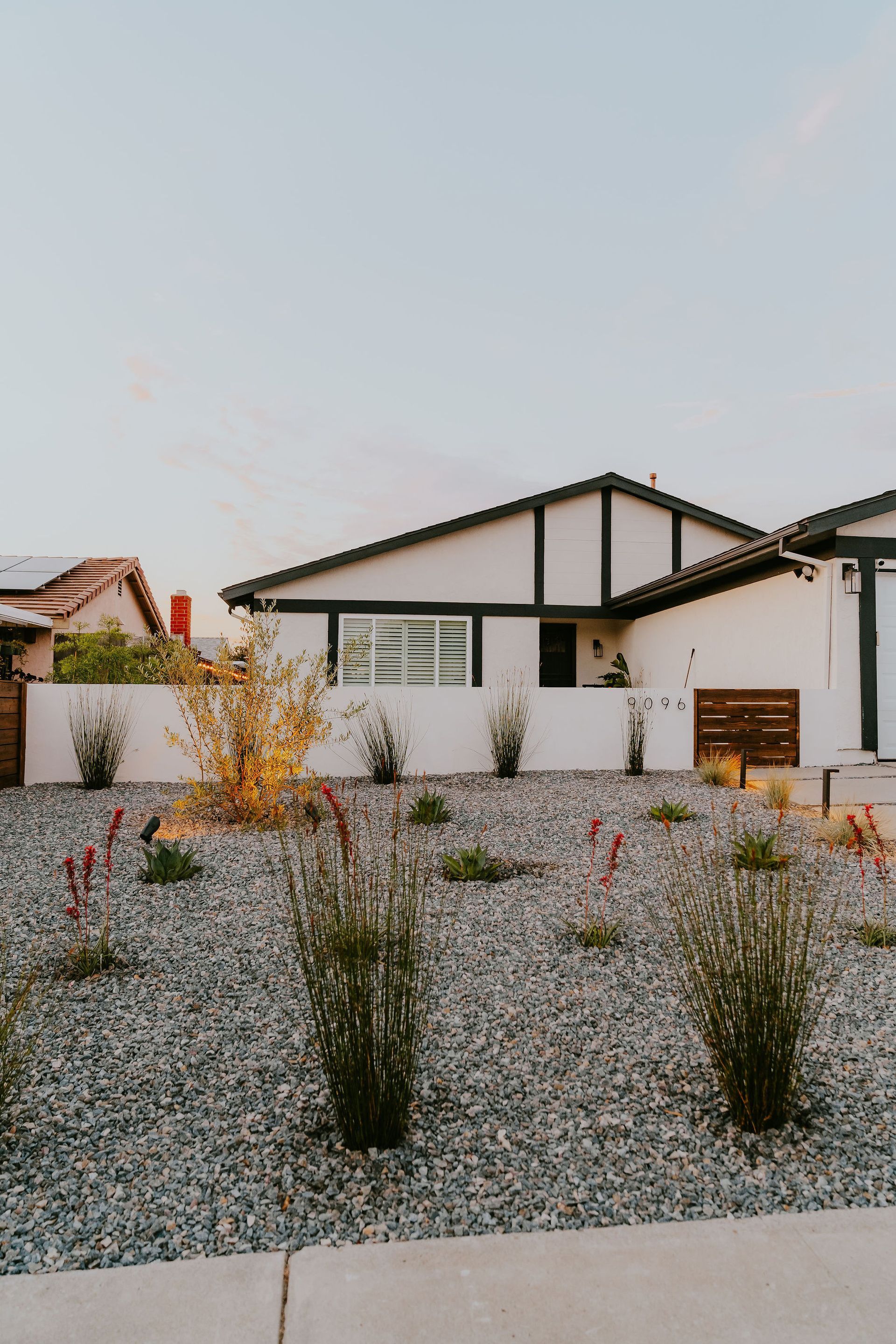 A modern, white single-story home with dark trim sits behind a gravel yard with scattered desert shrubs at dusk.