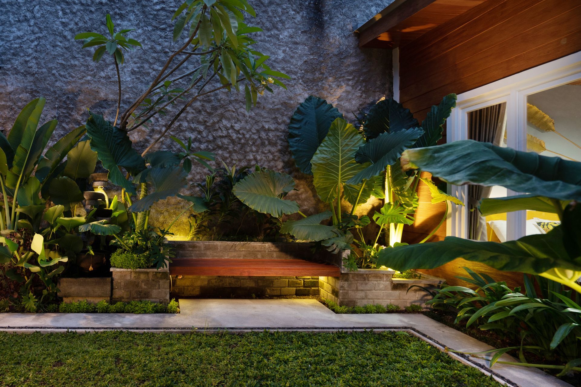 Lush, illuminated backyard garden with stone wall, bench, and tropical plants. Green grass, warm lighting, and wood trim.