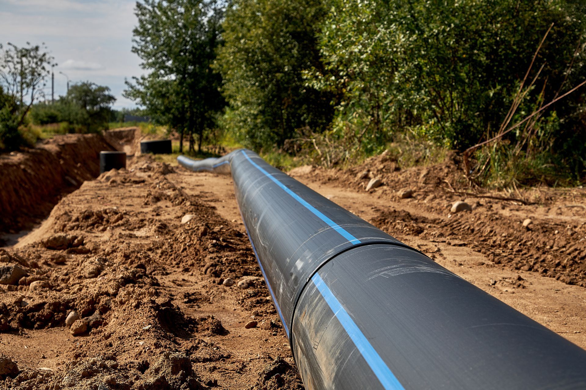 Black pipeline in a trench through a dirt area, with trees and a blue sky in the background.