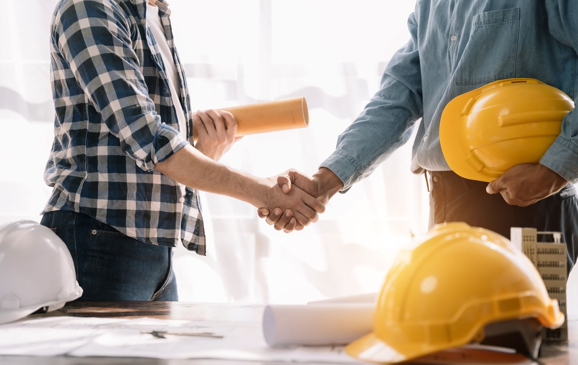 Two people shaking hands over a table with blueprints, hard hats, and a model building.