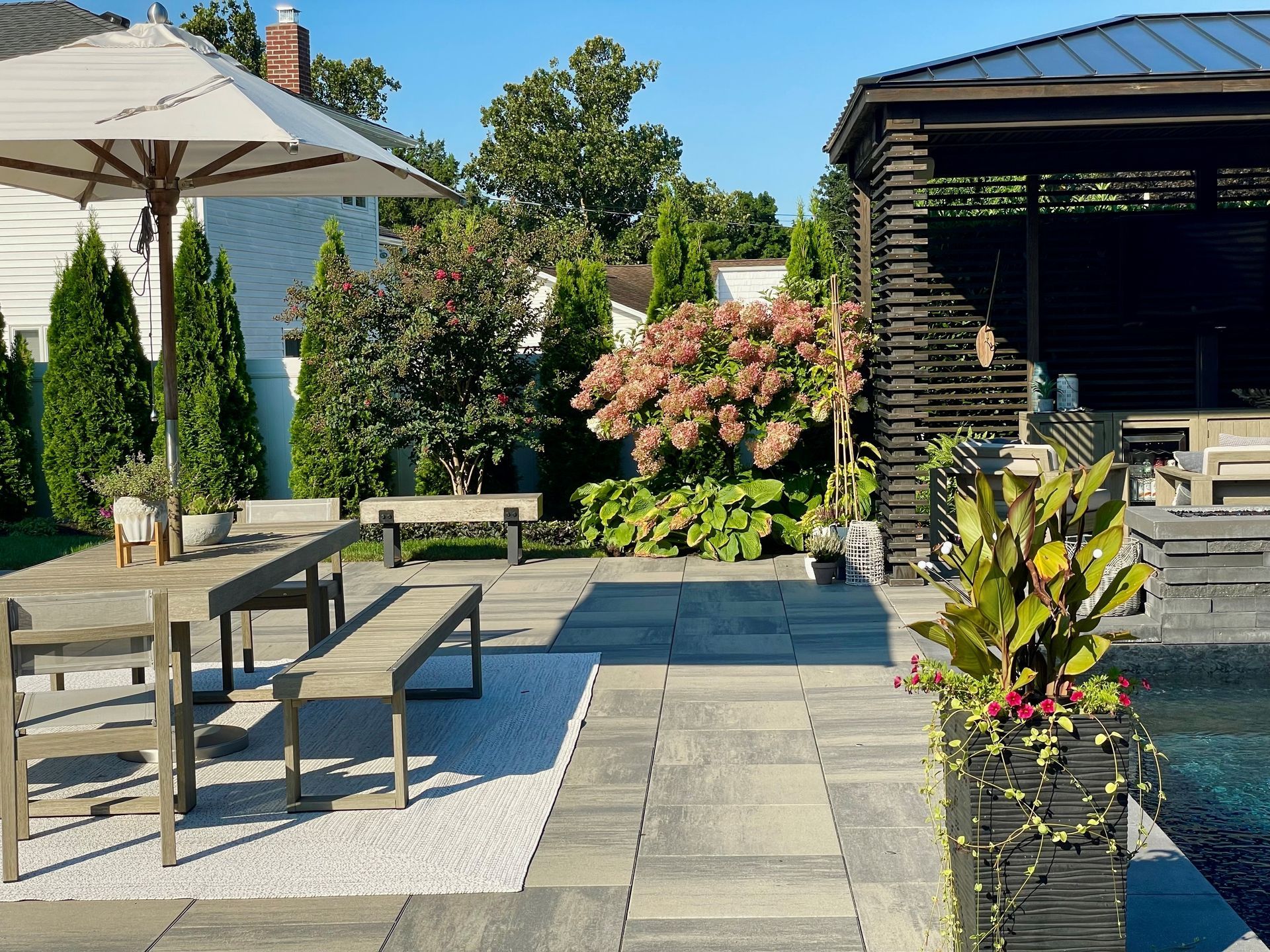 Patio with stone pavers, seating, an umbrella, and a wooden gazebo. Sunny day with lush greenery.
