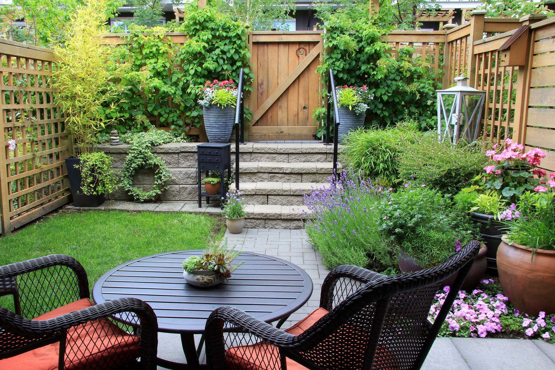 Cozy backyard patio with round table, wicker chairs, lush greenery, and wooden gate leading to a garden.