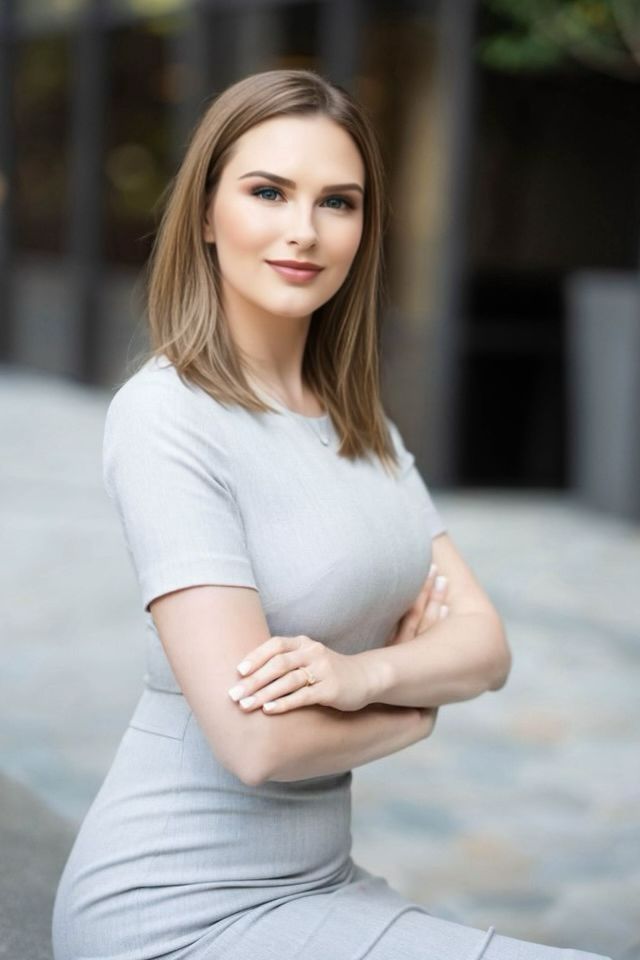 Blonde woman with crossed arms in grey dress smiles confidently outside a building.