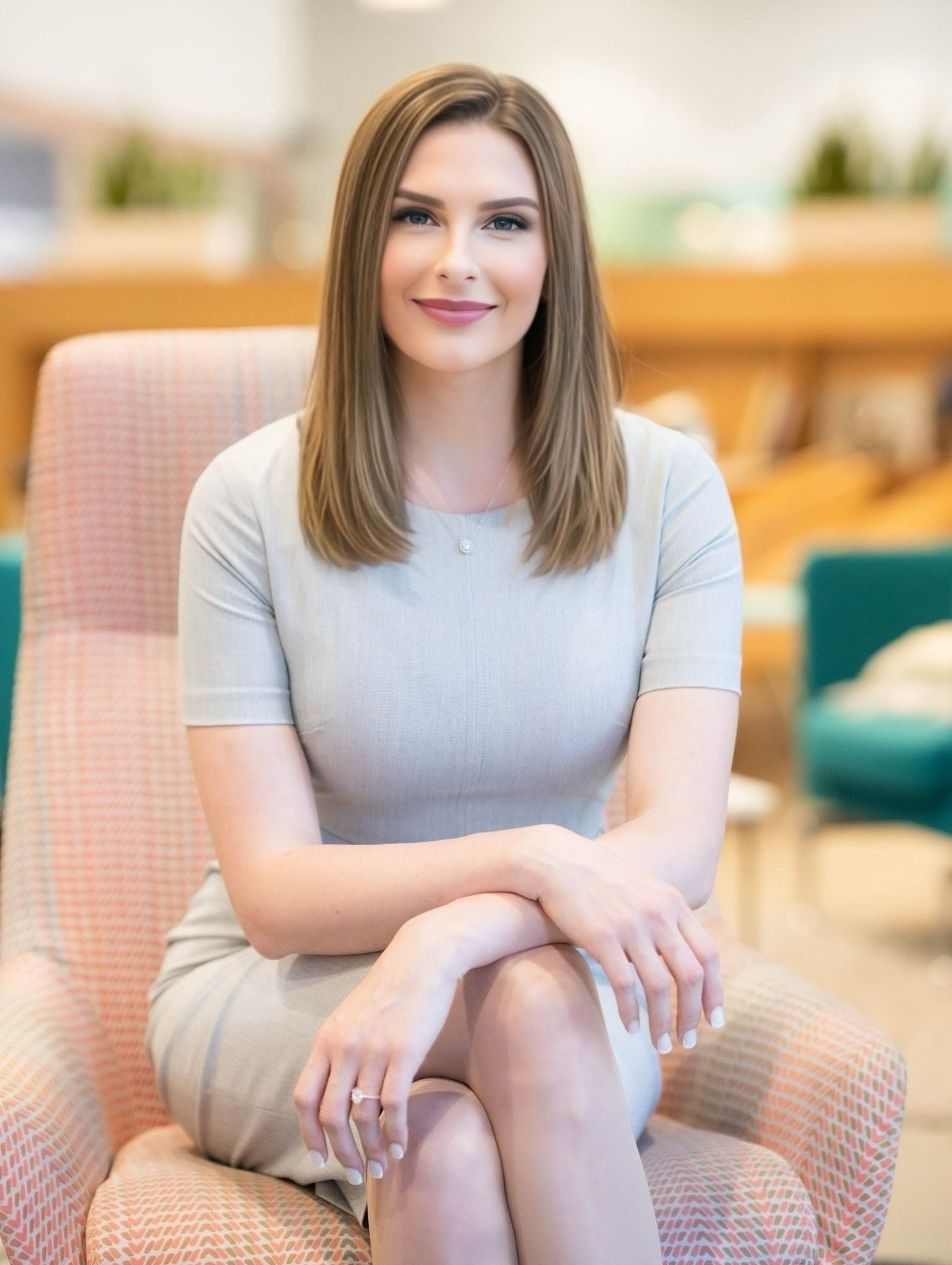 Woman in gray dress sits in pink chair, arms crossed, smiling. Modern office setting.