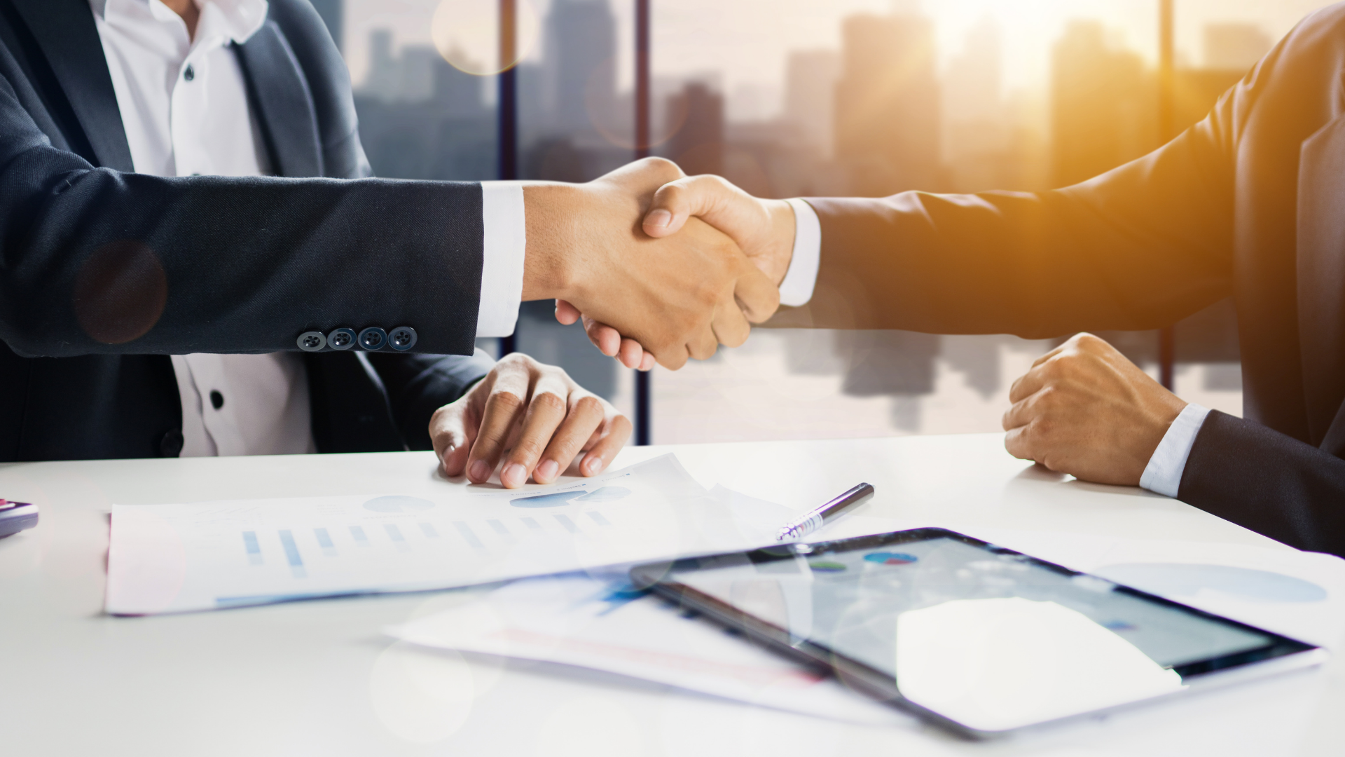 Businesspeople shaking hands across a desk with documents and a tablet in a bright office